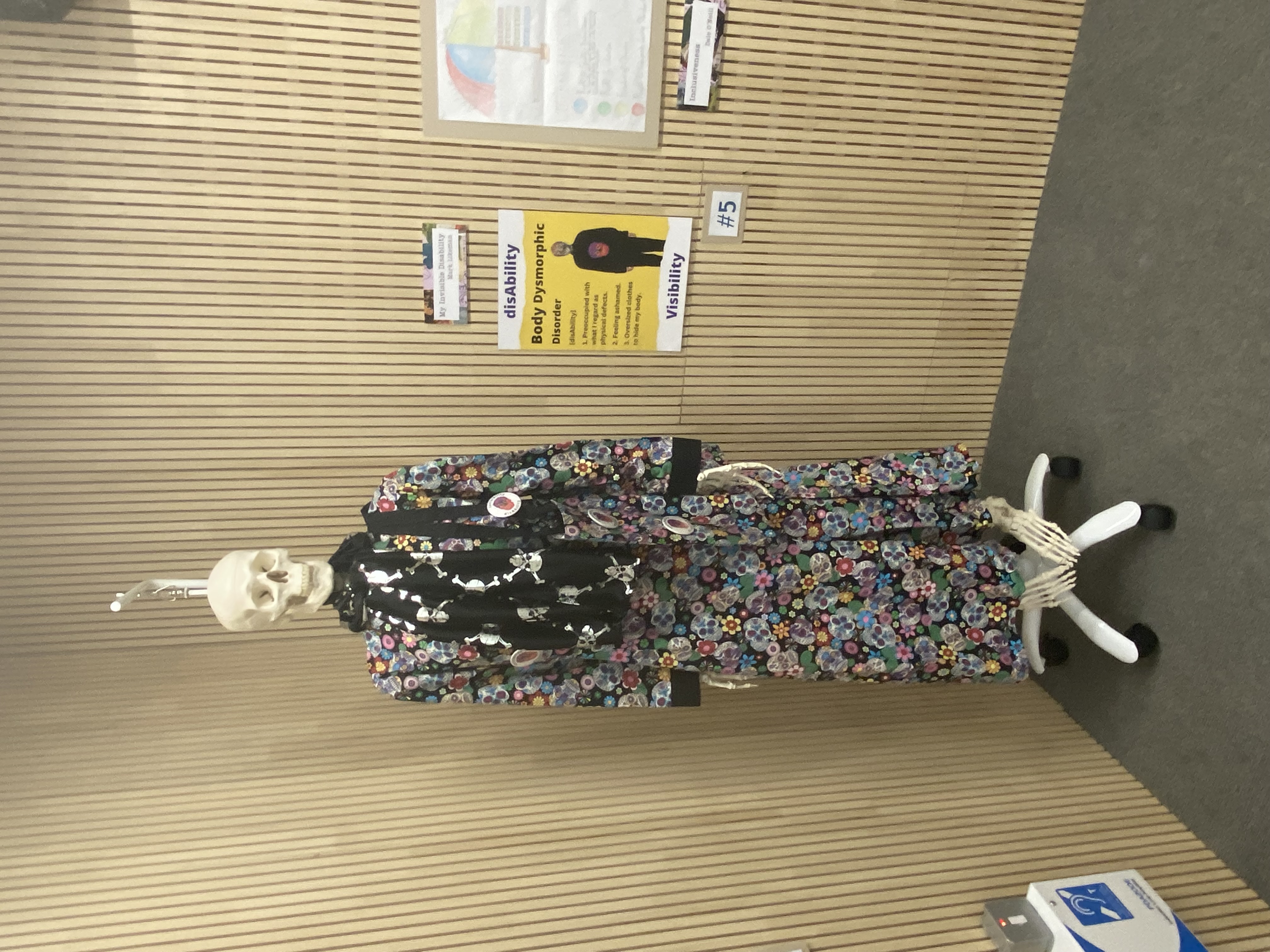 In this photo there is a human non-gender skeleton hanging on a stand with wheels. The skeleton is draped with a full-length dress, with a pattern of different coloured flowers and skulls. The are strips of black fabric (bias) on the neckline and cuffs of the dress. Around the neck of the skeleton is a long black scarf with silver skull and crossbones. There is a tote bag around the left shoulder of the skeleton, made of the same fabric as the dress. There are a number of badges or pins on the skeleton and tote bag that reads 'Invisibility disAbility', with a image of a skull adorned with different coloured flowers. The inspiration for the dress comes from The Day of the Dead (Día de Muertos or Día de los Muertos) a holiday traditionally celebrated on November 1 and 2, and widely observed in Mexico. The dress is made from 100% cotton twill fabric. To the right of the skeleton there is a poster the reads 'disAbility Invisibility'. Body Dysmorphic Disorder. Below this are the words; 1. Preoccupied with what I regard as physical defects. 2. Feeling ashamed. 3. Oversized clothes to hide my body. There is also an image of a male figure with face covered and wearing a boiler suit, with a skull on the front.
